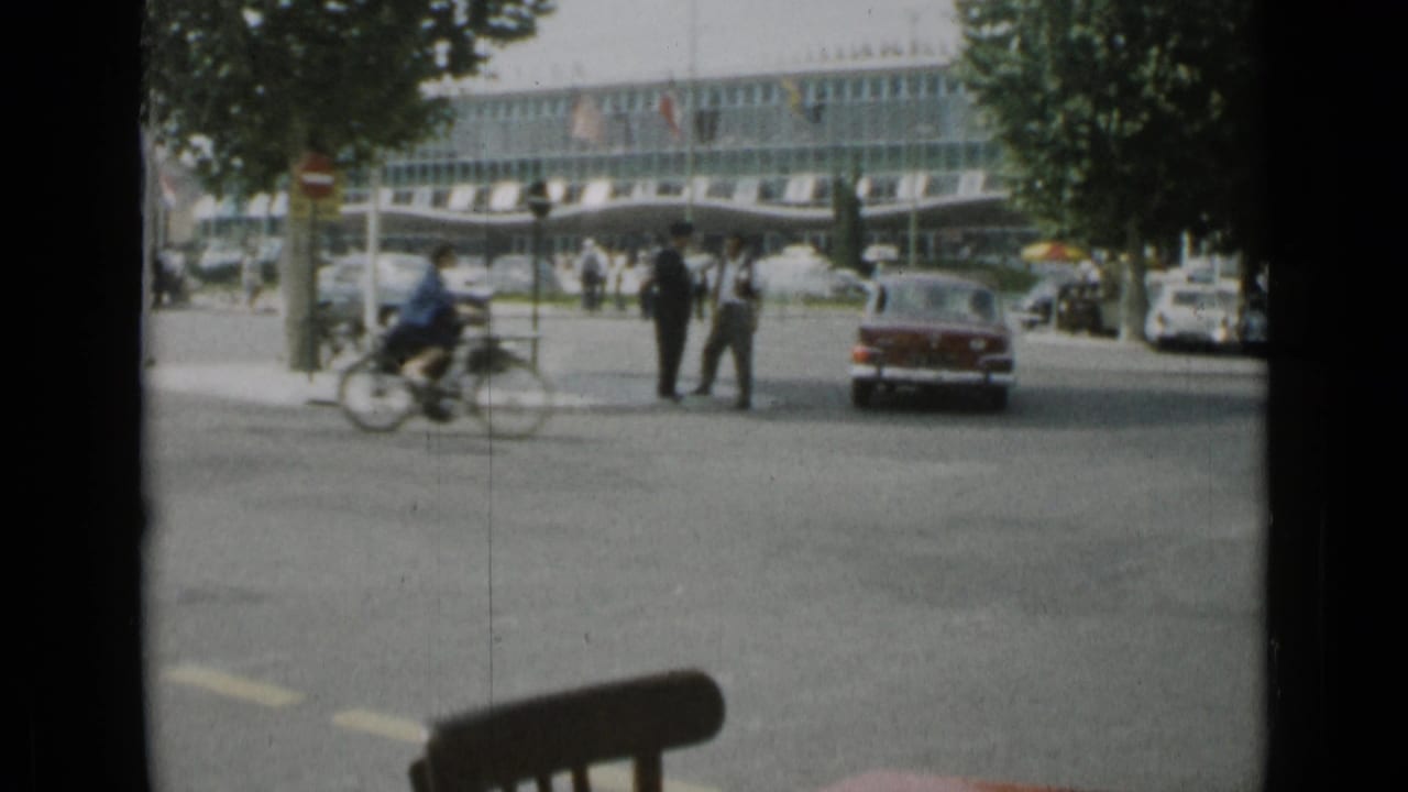 Street intersection with red sedan, pedestrians, cyclist, trees, and glass-fronted building.