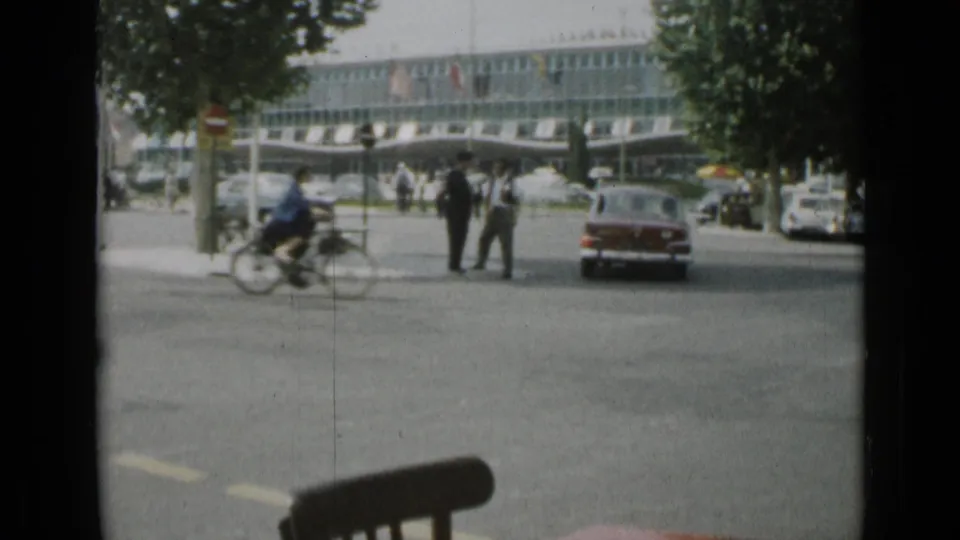 1962 Cannes street view through a dark window edge, with a red sedan, cyclist, pedestrians, trees, and a modern glass-fronted building.