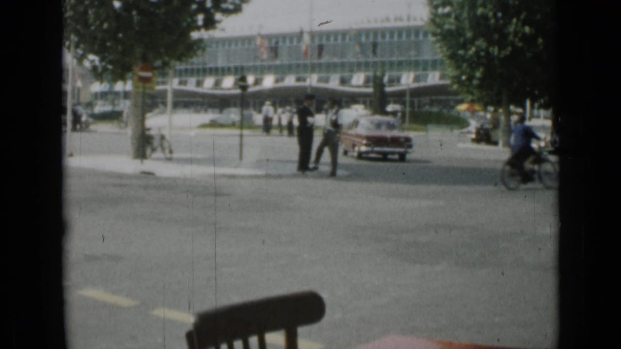 Intersection with pedestrians, red car, motorbike or cyclist, trees, and commercial building.