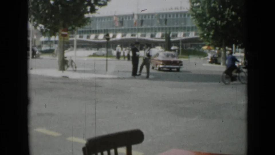Cannes street scene from 1962 showing pedestrians near an intersection, a red car, a passing rider, trees, and a glass commercial facade.