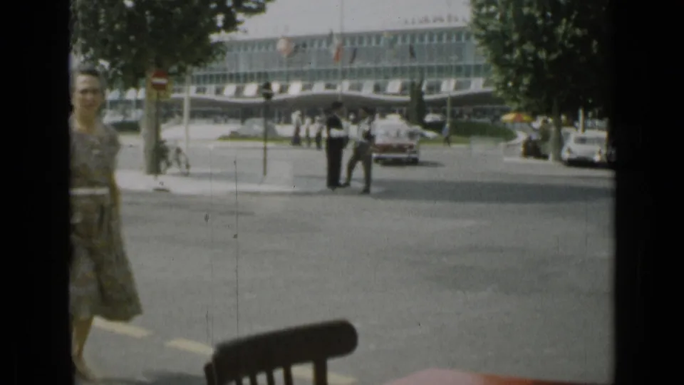 1962 Cannes street view with a woman partly entering the left edge, pedestrians in the distance, a red car, trees, and a glass-fronted building.