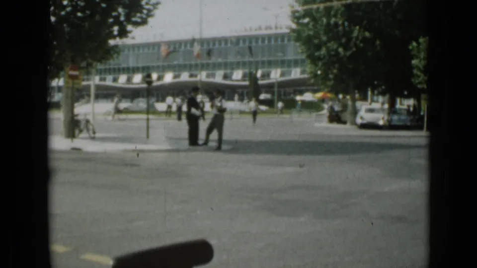 Cannes street corner in 1962 with pedestrians gathered near the roadway, parked cars under trees, and a modern glass-fronted commercial building.