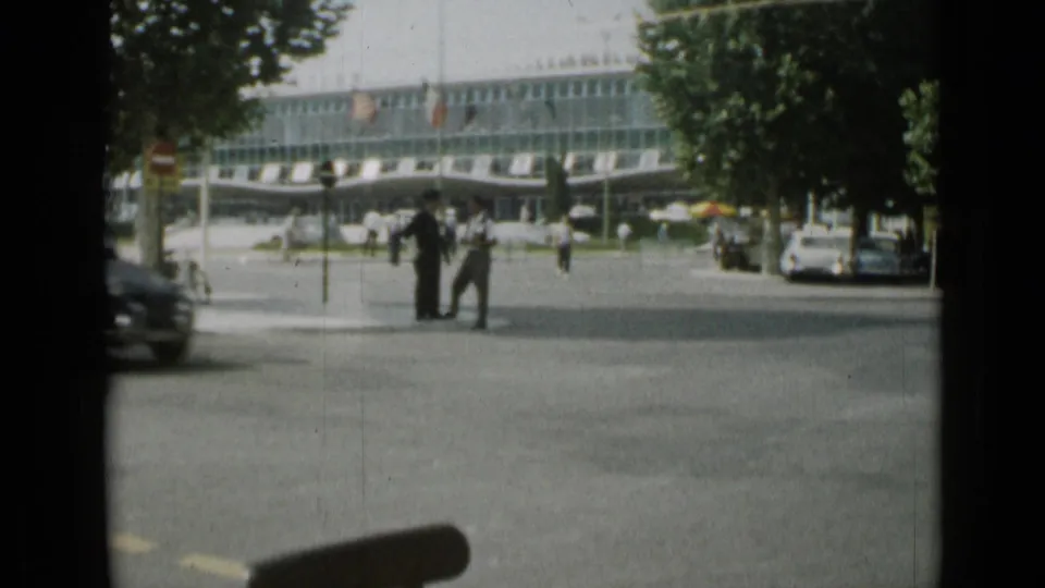 1962 Cannes street view showing pedestrians near a modern building, with trees, traffic, and the edge of an 8mm frame visible.