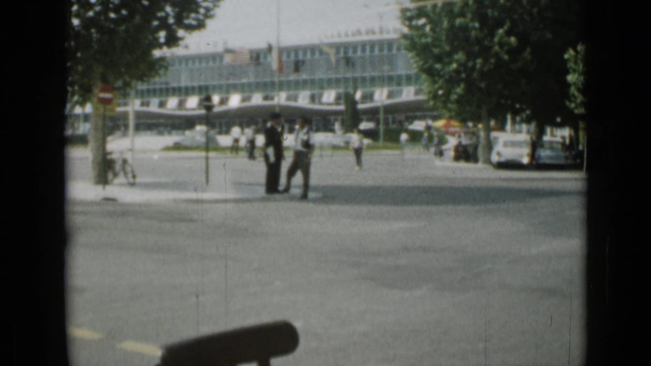 Pedestrians near a street crossing with a modern building and cars in the background.