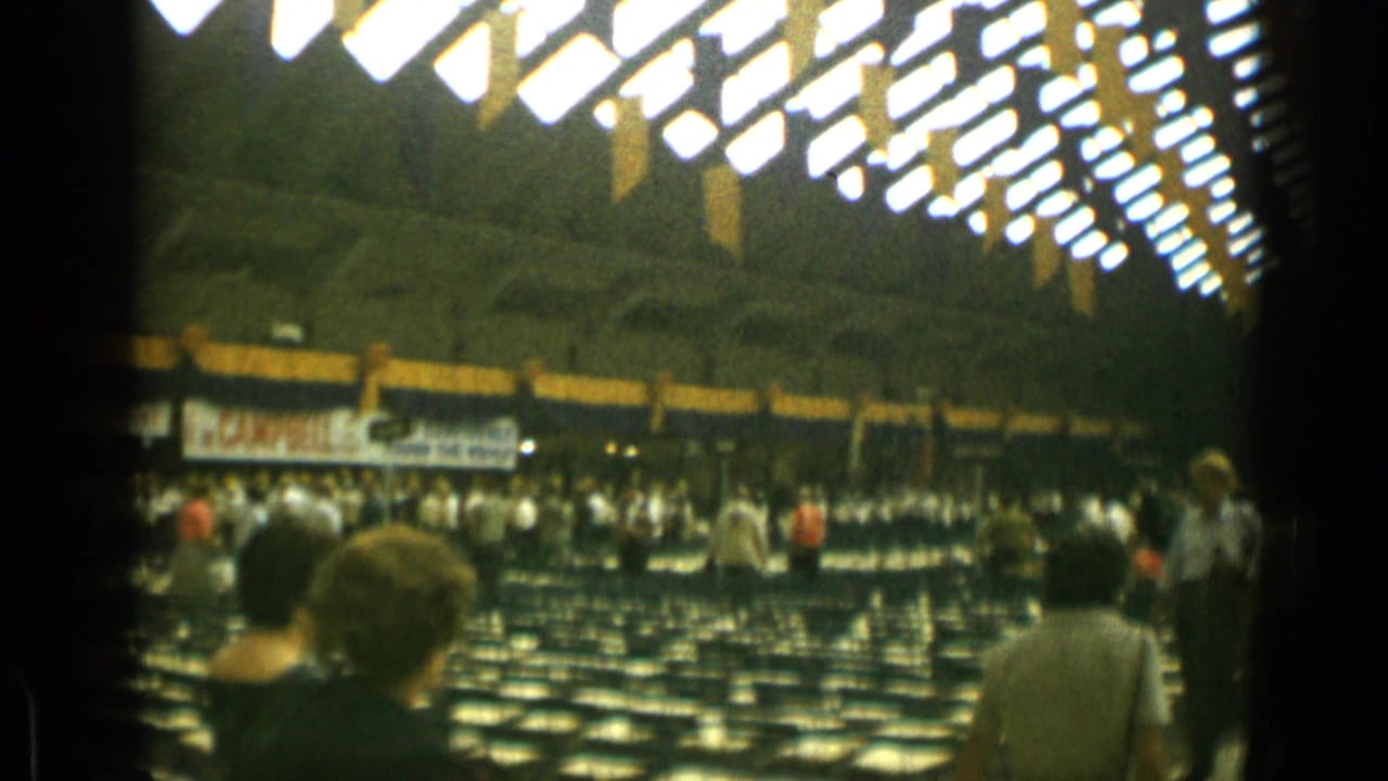 Crowd moving through rows of empty chairs under bright auditorium ceiling lights.