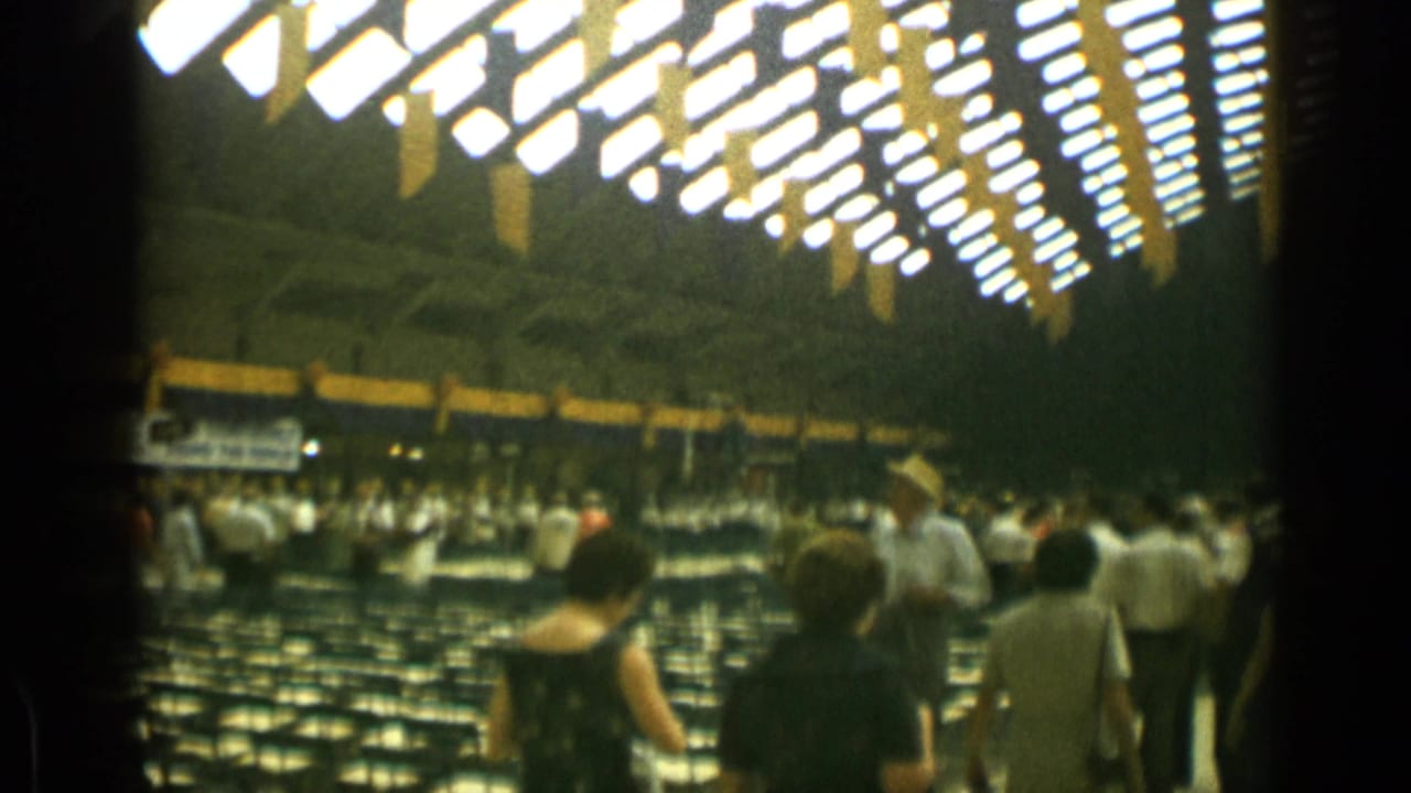 People walk beside rows of chairs in a large hall with patterned ceiling lights.