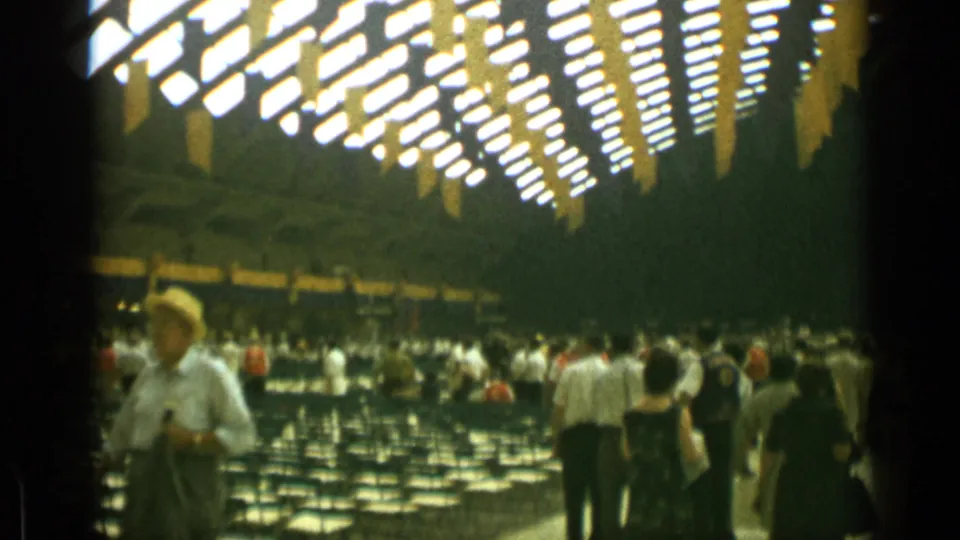 Handheld 8mm view of people walking away through a Cannes auditorium with chair rows and bright ceiling panels overhead.