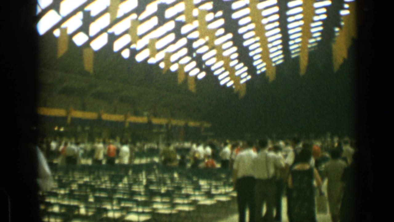Crowd inside a large hall with rows of chairs and bright ceiling lights.