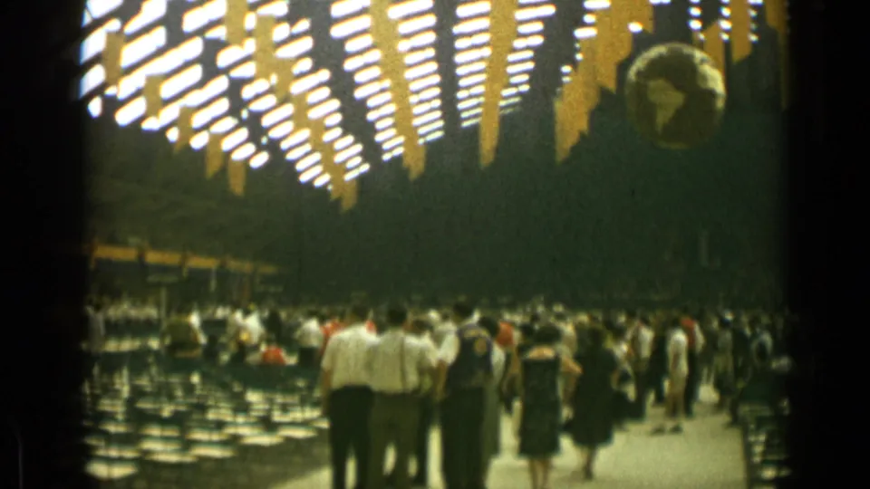 1962 Cannes home-movie frame showing adults in mid-century clothing gathered beneath bright ceiling lights and yellow hanging decorations.