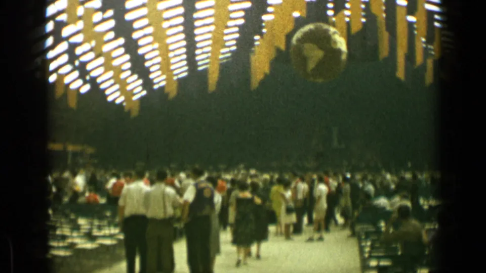 1962 Cannes frame showing a busy indoor hall with adults walking, rows of seating, yellow ceiling decorations, and a suspended globe.