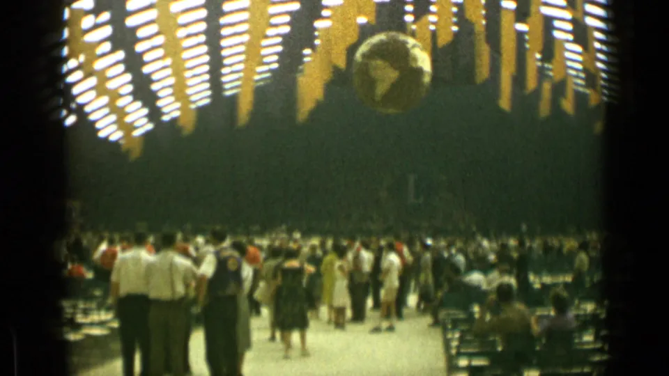 1962 Cannes home-movie frame showing attendees in a large decorated hall with rows of chairs, ceiling lights, and a suspended globe.