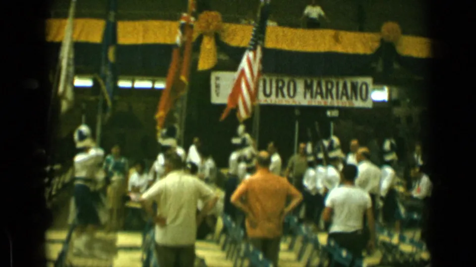 1962 Cannes home-movie frame with men watching activity near multiple flags beneath a partially visible Arturo Mariano banner.