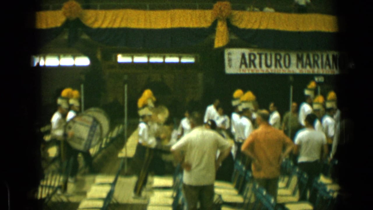 Marching band in white uniforms performs before seated spectators inside a decorated venue.