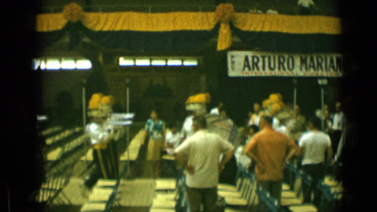 Band members with drums and brass instruments move past spectators under yellow-and-black decorations.