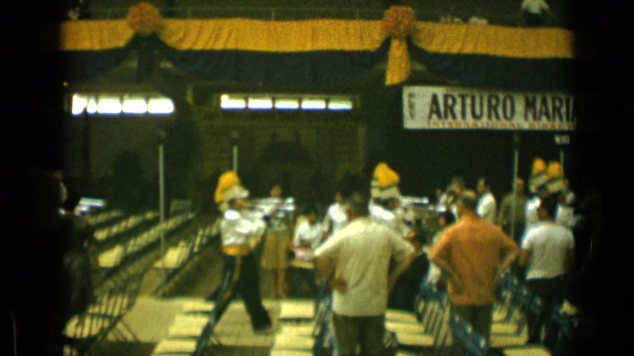 Marching band members in white uniforms perform before spectators in a decorated indoor hall.