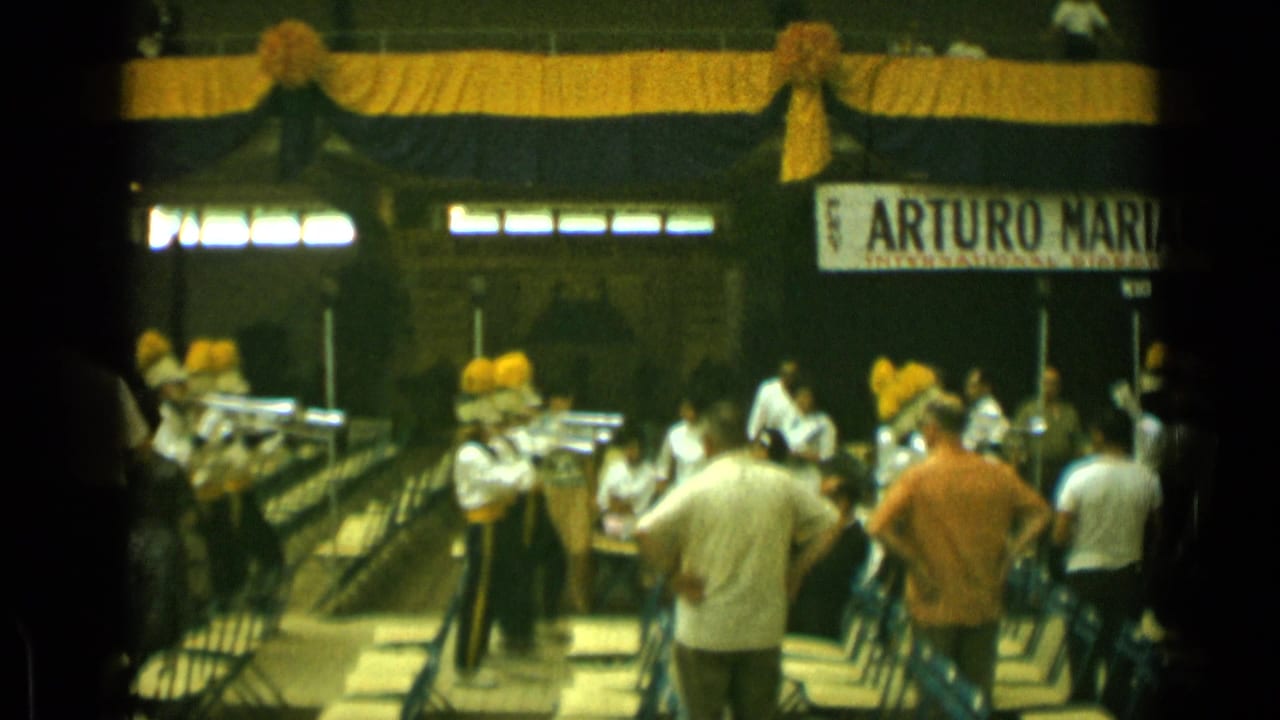Uniformed band members and spectators stand near rows of folding chairs under a stage sign.