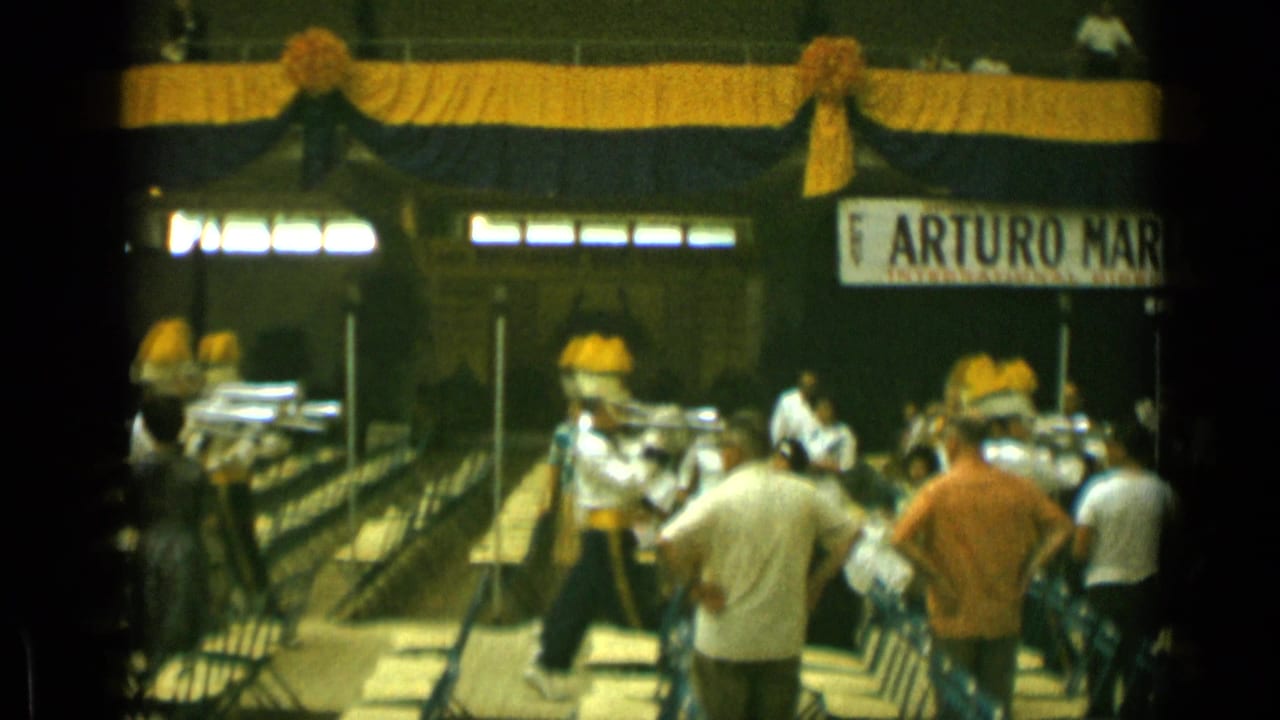 Marching band members in white uniforms perform before seated spectators under draped decorations.