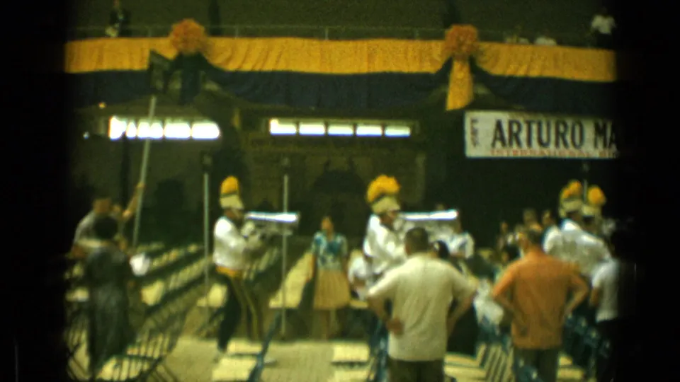 Cannes public-event footage shows spectators and uniformed musicians beneath bold drapery, with a partial sign visible in the background.