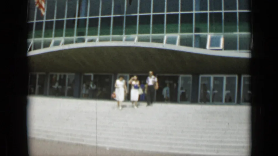 Exterior Cannes frame showing adults leaving or crossing a plaza in front of a modernist building with large glass windows and doors.