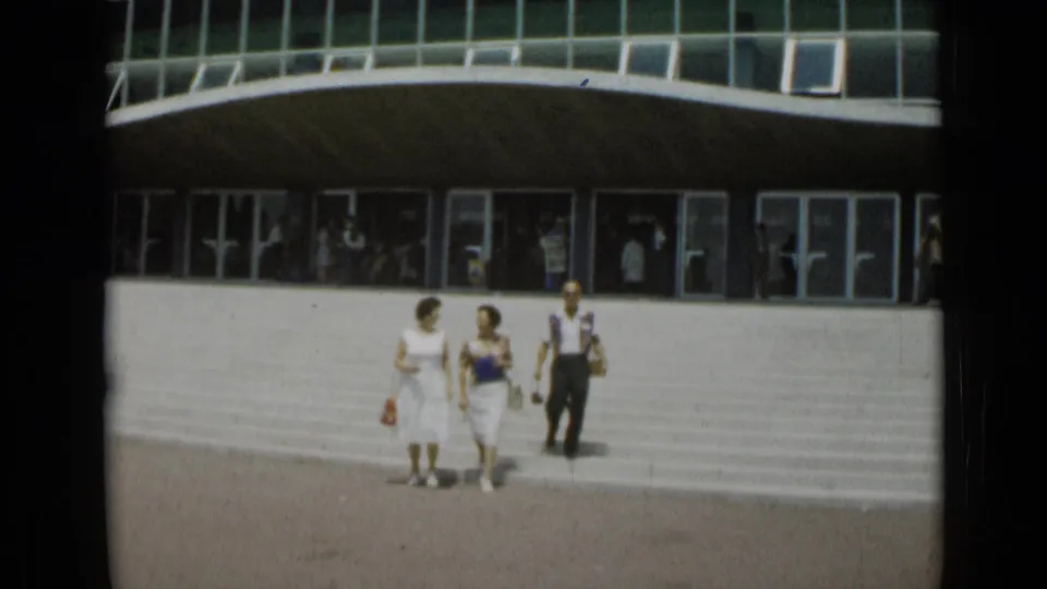 In Cannes, France in 1962, two women and one man walk toward the camera across a broad concrete plaza outside a modernist building.