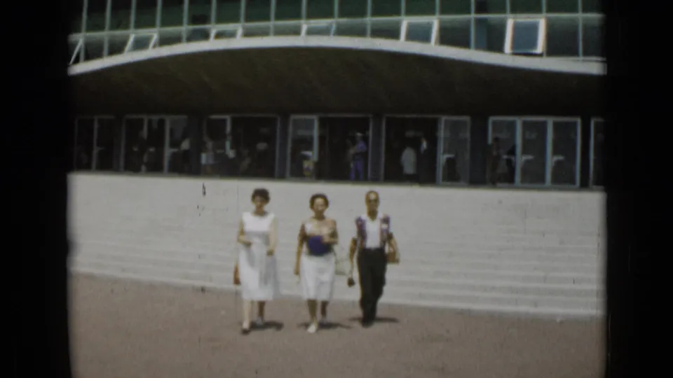 The three adults continue walking toward the camera in front of a curved glass-and-concrete facade in 1962 Cannes.