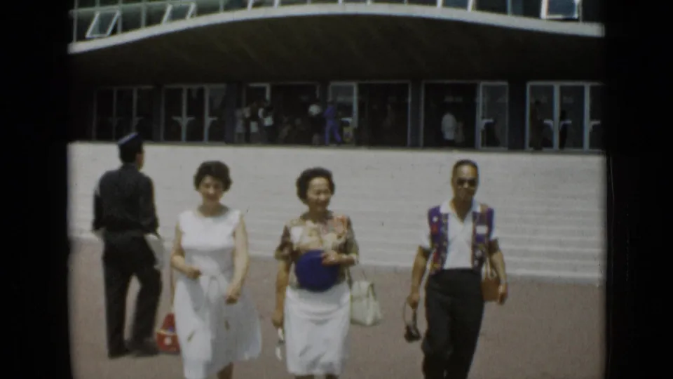 In Cannes, France in 1962, two women and a man walk toward the camera across a bright parking area outside a modern building.