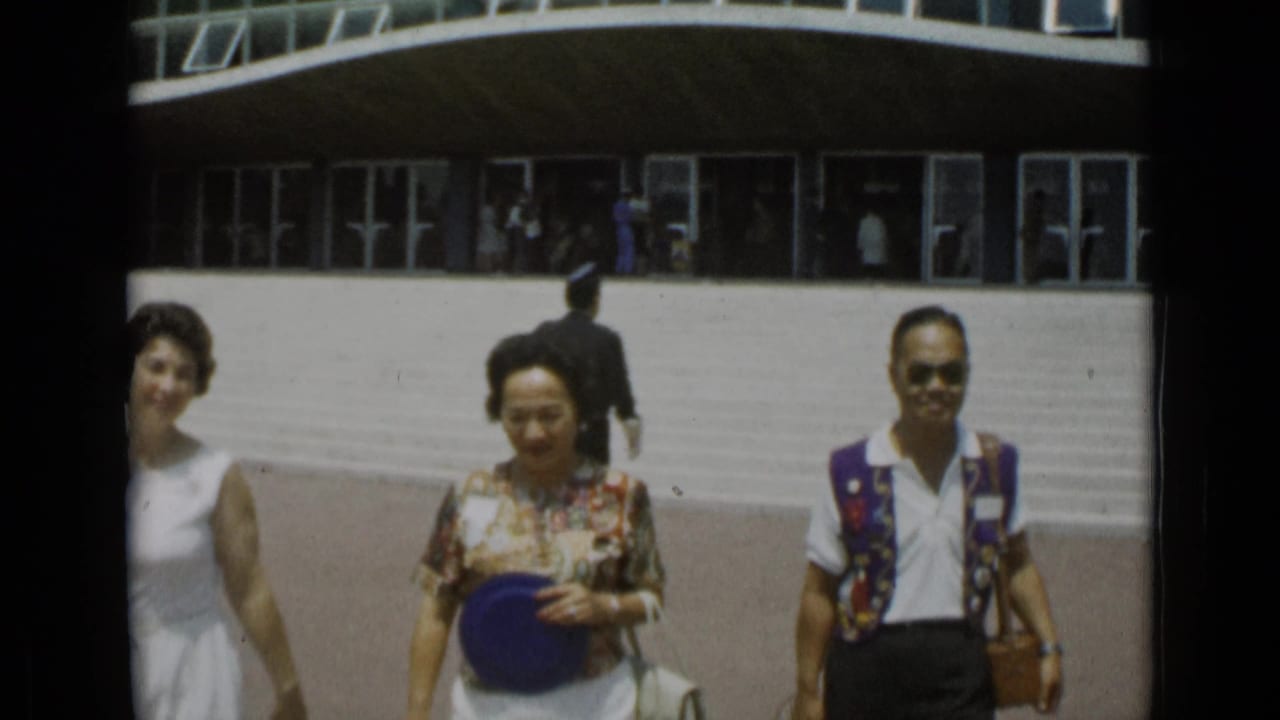 Two women and a man in 1960s clothing approach the camera in a Cannes parking area, with steps and glass doors behind them.