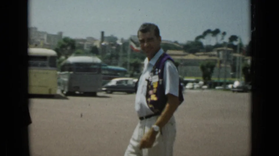 The man walks across the Cannes parking area as buses, cars, low buildings, and hillside structures appear in the background.