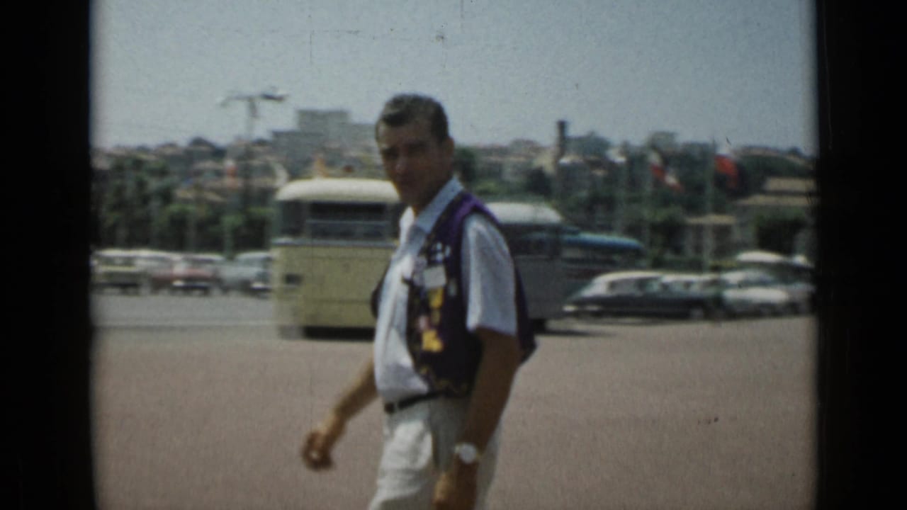 Man in a white shirt and dark vest walking across a sunny street with buses and cars behind him.