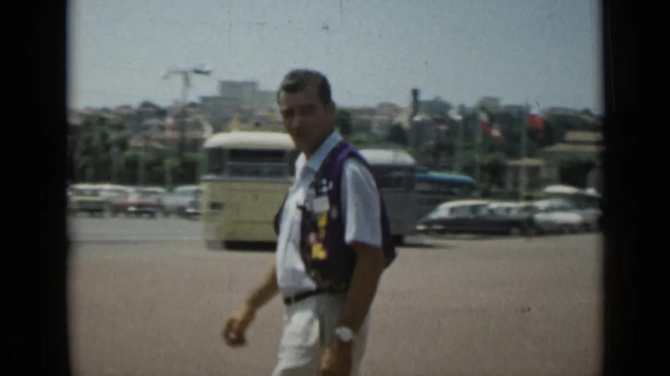 In Cannes, France, in 1962, a man in a white shirt and dark vest walks past the camera on a bright street with buses, cars, and palm trees behind him.
