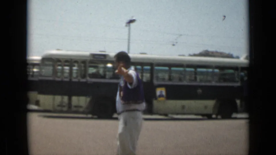 In the 1962 Cannes home-movie scene, the man raises a hand toward the camera while a long bus fills the background.
