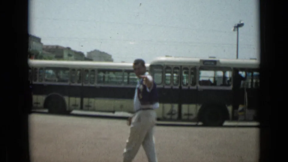 The man waves toward the camera in Cannes, France, in 1962, standing against a background of a street bus and distant buildings.