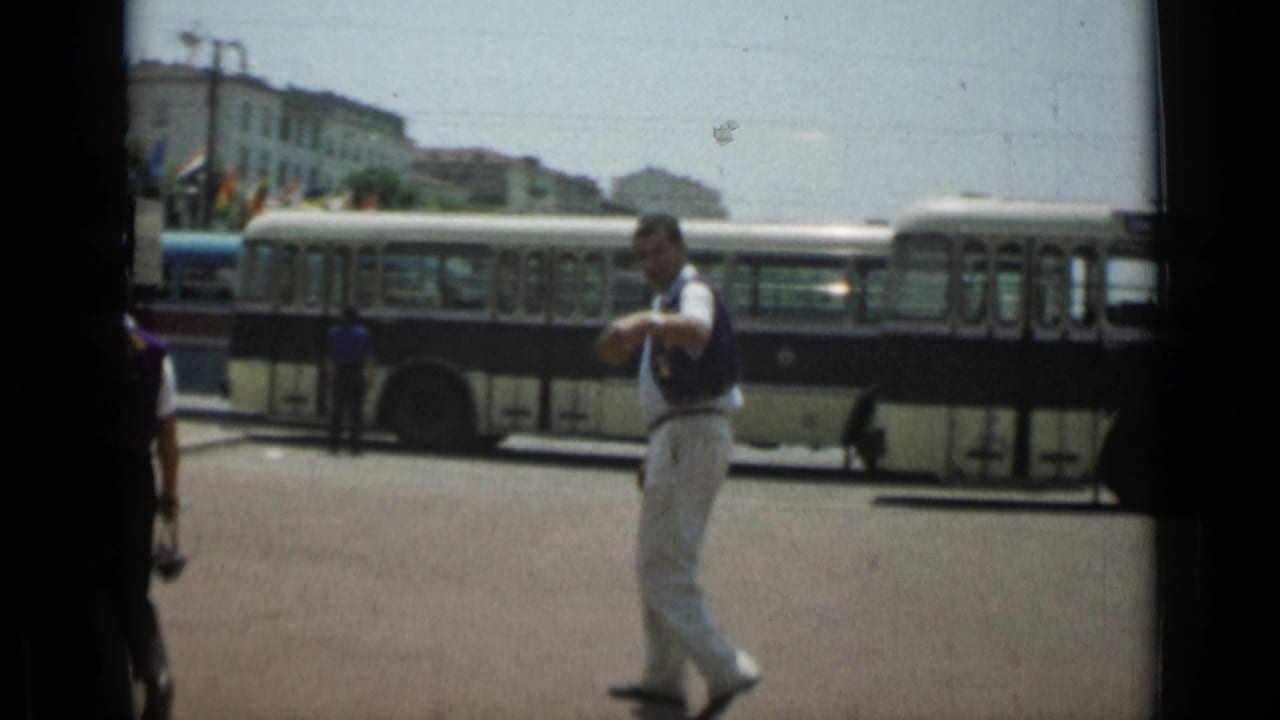 Man in white trousers walks in front of a long bus on a sunny city street.