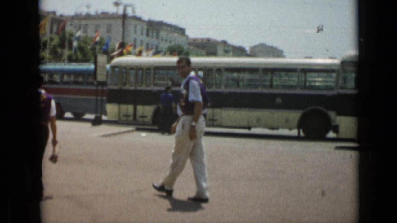 Man carrying a small item walks past a bus with flags and buildings behind him.