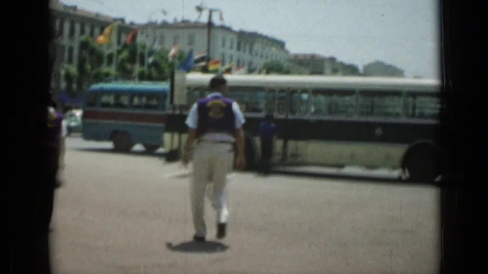 A 1962 Cannes street frame follows the man from behind as he approaches the bus, with flags and urban architecture across the road.