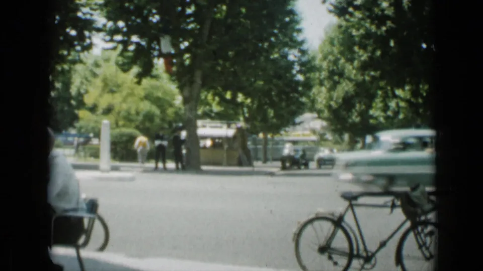 The closing 1962 Cannes frame shifts to a tree-lined street with a bicycle at right, passing cars, and pedestrians near the roadside.
