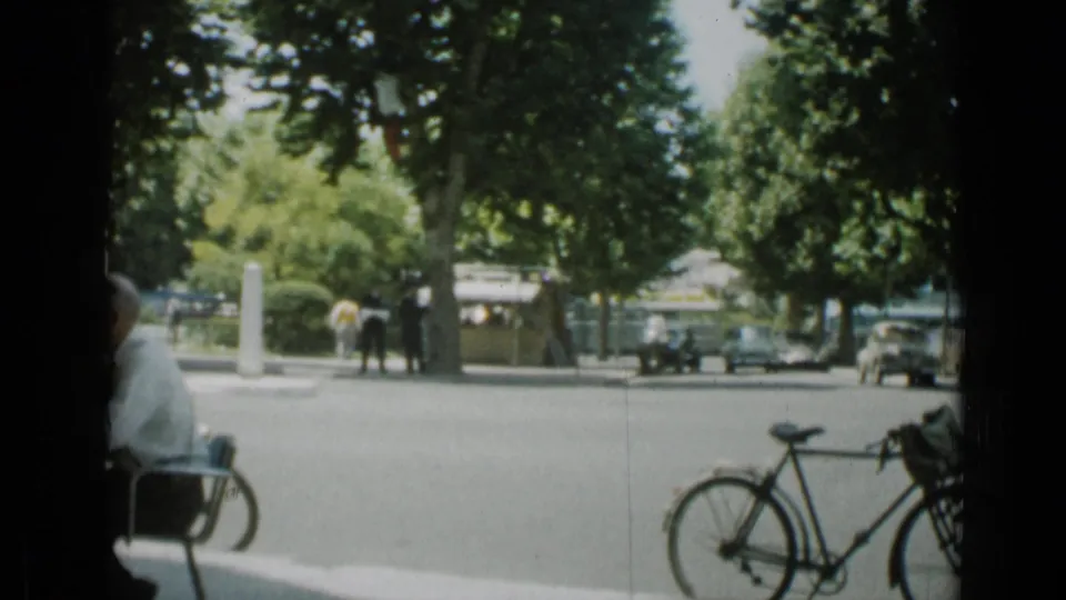 1962 Cannes street view with a seated man in shade, a bicycle in the foreground, pedestrians, trees, and parked cars beyond.