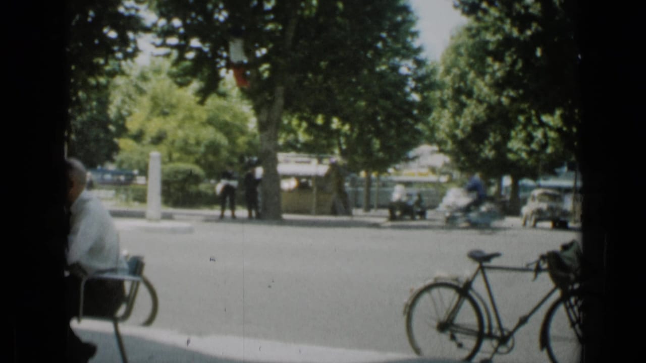 Cannes street in 1962 with a bicycle near the camera, pedestrians across the road, trees, parked cars, and a blurred motorbike passing.