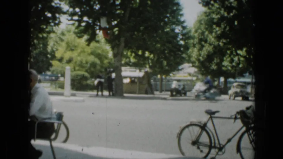 Cannes street in 1962 with a bicycle near the camera, pedestrians across the road, trees, parked cars, and a blurred motorbike passing.