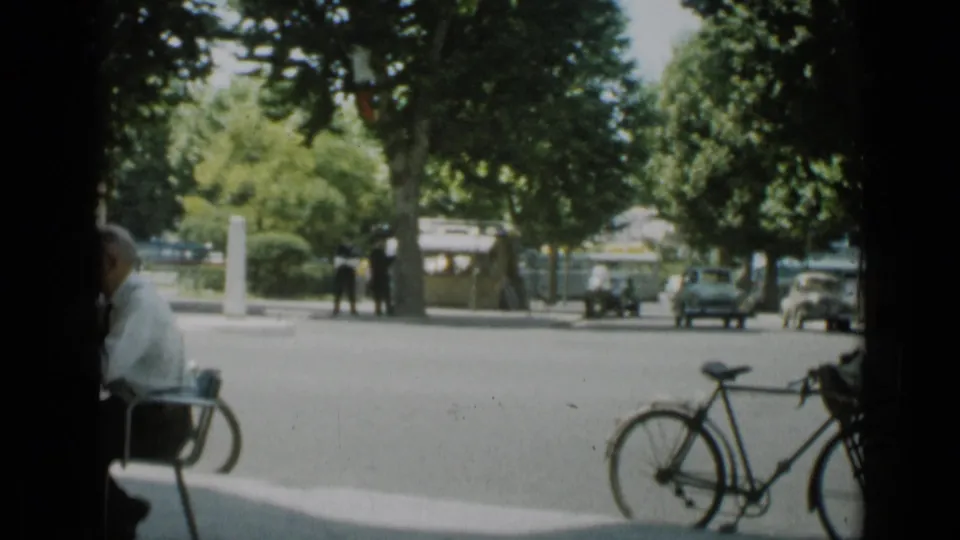 A 1962 Cannes sidewalk scene shows a seated man at left, bicycles in the foreground, pedestrians, leafy trees, and classic cars across the road.
