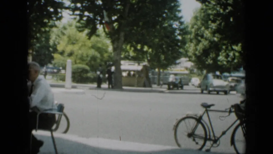 End frame of the 1962 Cannes street view, with a bicycle foreground, seated man in shade, pedestrians near trees, and a light-colored car moving or parked.