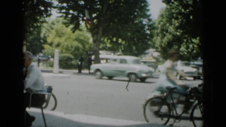 1962 Cannes street view from an outdoor restaurant, with vintage cars passing, bicycles near the curb, and cafe seating in the foreground.