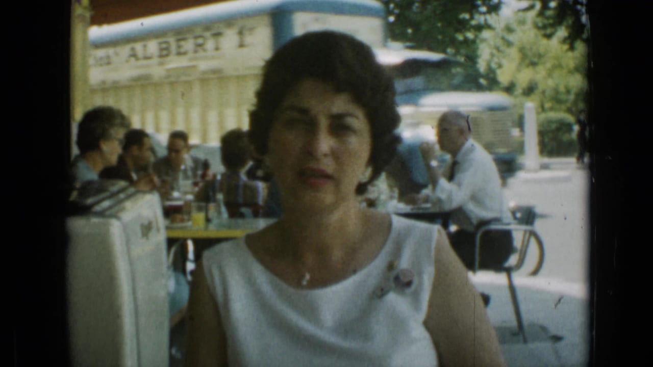 Woman in a white sleeveless dress seated at an outdoor cafe with diners behind her.