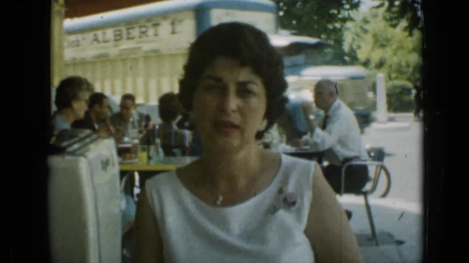A woman sits at an outdoor restaurant in Cannes in 1962, with cafe patrons, drinks, a partial sign, and traffic visible behind her.