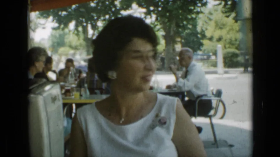 The seated woman turns away from the camera at a shaded Cannes outdoor restaurant, with other patrons and a tree-lined street in the background.