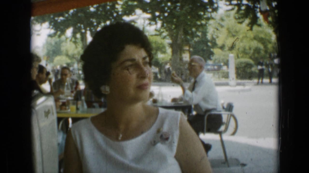 Woman in a white sleeveless dress seated at an outdoor cafe with patrons behind her.