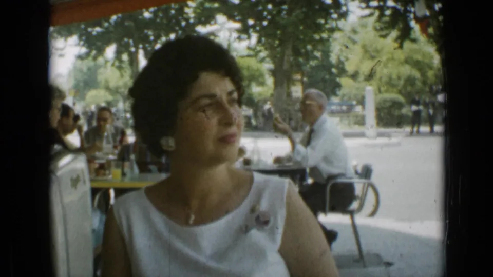 Adult woman at a shaded outdoor cafe in Cannes, France, with other patrons seated nearby in 1962 home-movie footage.