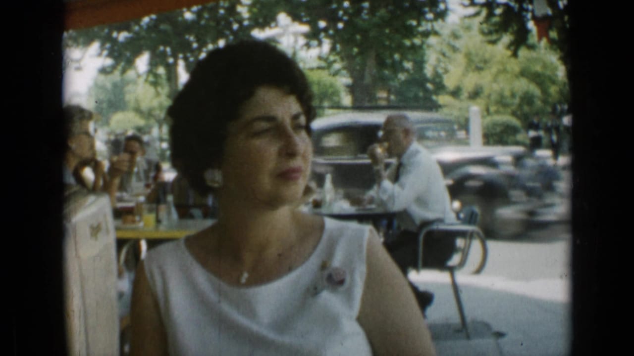 Woman seated at an outdoor cafe as a dark vintage car passes in the background.