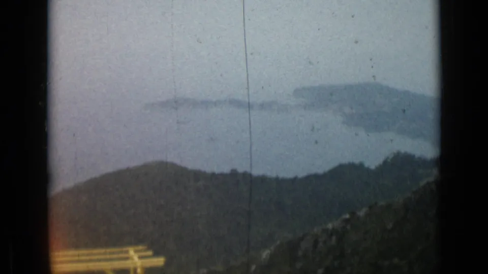 Brief 8mm landscape cutaway showing a hazy mountain view with trees and slopes, following the Cannes cafe scene.