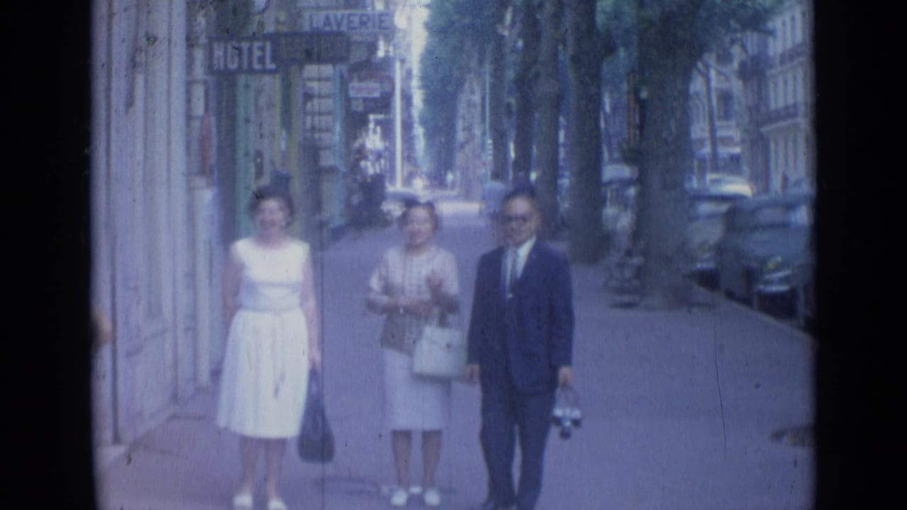 Three adults in 1960s dress walk toward the camera on a Cannes sidewalk with hotel and laverie signs behind them.